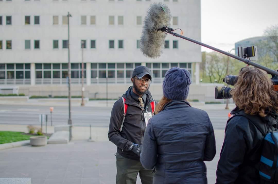 DaVante' Goins being interviewed on camera with a boom mic outside a government building